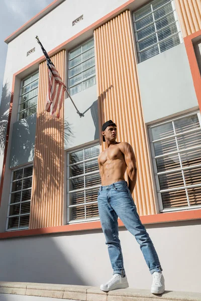 Shirtless cuban man in baseball cap and jeans standing on parapet near building with american flag — Stock Photo