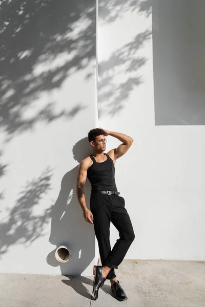 Joven hombre cubano con gafas y camiseta negra sin mangas posando cerca de la pared en Miami, durante el verano - foto de stock