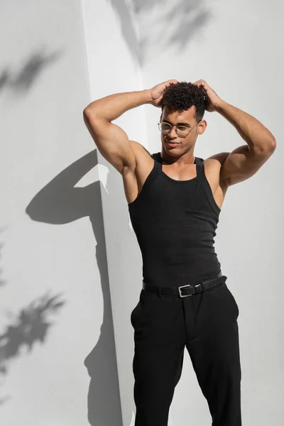 Rilassato muscolare giovane cubano in occhiali e senza maniche t-shirt toccare i capelli vicino alla costruzione — Foto stock