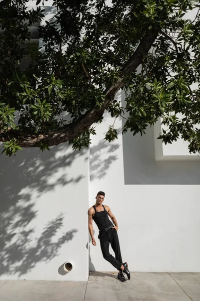Stylish young cuban man in black outfit and eyeglasses posing near building with shadow — Stock Photo