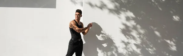Muscular cuban man in eyeglasses and black outfit looking away while standing near building, banner — Stock Photo