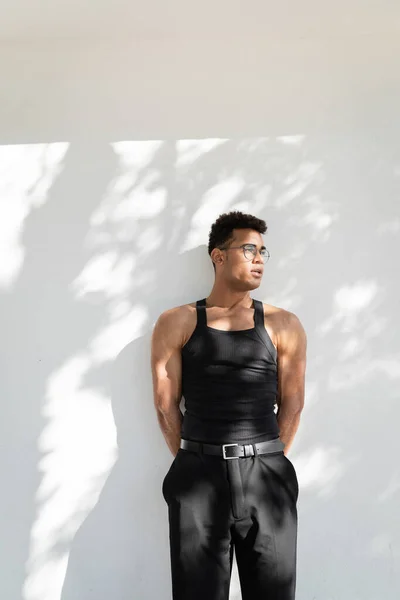 Joven y musculoso joven cubano con gafas y atuendo negro de moda mirando hacia otro lado - foto de stock