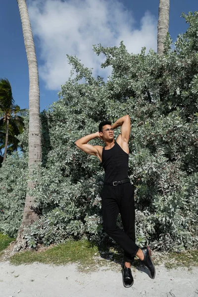 Hombre cubano musculoso en traje negro y anteojos cerca de plantas verdes al aire libre en Miami, playa sur - foto de stock