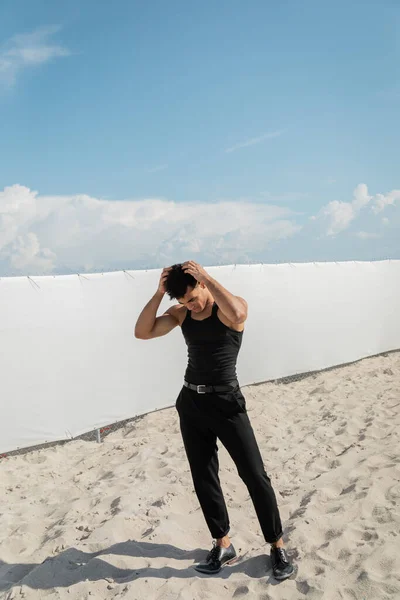 Homme cubain en tenue noire toucher les cheveux tout en se tenant sur le sable sur la plage à Miami — Photo de stock