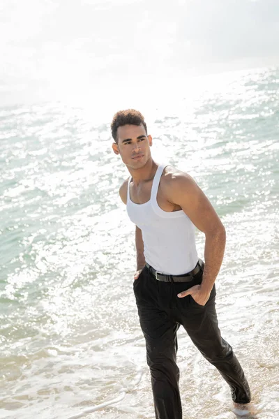 Hombre cubano cogido de la mano en bolsillos de pantalones mientras camina sobre arena con agua del océano en Miami - foto de stock