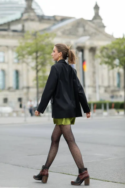 Vista lateral de la mujer joven con estilo en chaqueta negra y botas mirando hacia otro lado mientras camina en Berlín - foto de stock