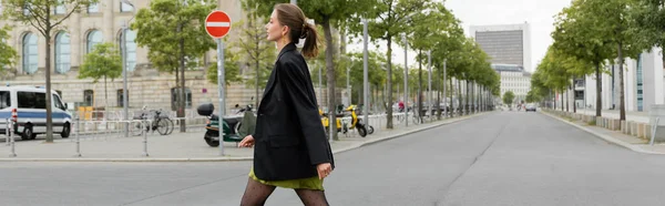 Seitenansicht einer eleganten und stilvollen jungen Frau in Kleid und schwarzer Jacke, die in Berlin unterwegs ist, Banner — Stockfoto
