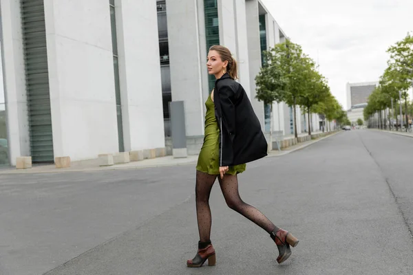 Alla moda giovane bella donna in giacca e abito di seta verde guardando lontano mentre cammina sulla strada — Foto stock