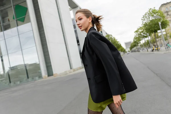Smiling young and fair haired woman in black blazer, silk dress looking away while walking in Berlin — Stock Photo