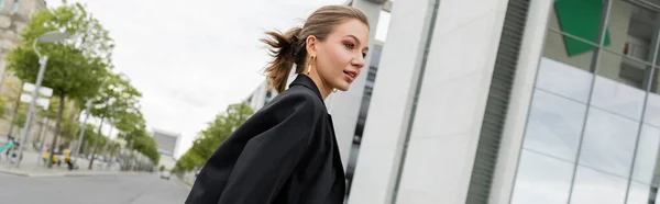 Positive young fair haired woman in black blazer looking away while walking in Berlin, banner — Stock Photo
