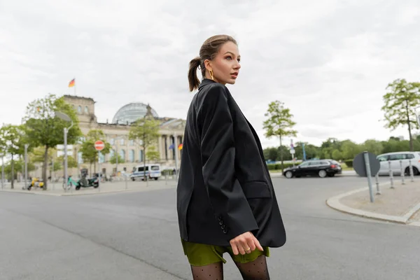 Joven mujer de pelo justo con estilo en chaqueta negra y vestido mirando hacia otro lado mientras camina en Berlín - foto de stock
