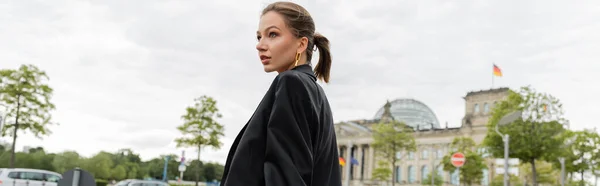 Giovane donna alla moda dai capelli biondi in giacca nera guardando altrove mentre in piedi a Berlino, striscione — Foto stock