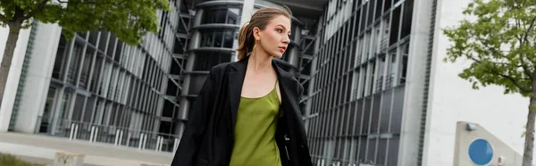 Giovane donna dai capelli biondi in abito di seta verde e blazer che cammina a Berlino, Germania, banner — Foto stock