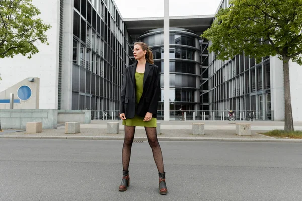 Pleine longueur de jeune femme branchée en bottes et blazer élégant regardant loin dans la rue à Berlin — Photo de stock