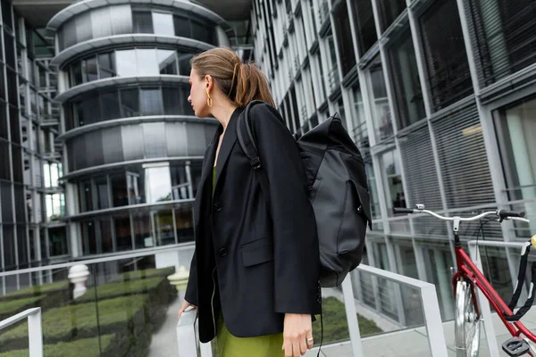 Side view of stylish fair haired woman in black jacket and backpack standing near bicycle — Stock Photo