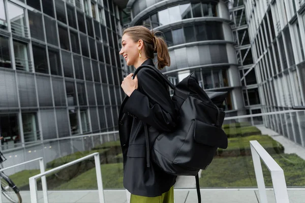 Side view of cheerful and stylish young woman in black jacket holding backpack and looking away — Stock Photo