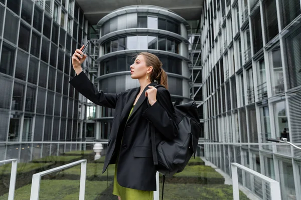 Вид збоку модна молода жінка в куртці тримає рюкзак і приймає селфі на смартфон — стокове фото
