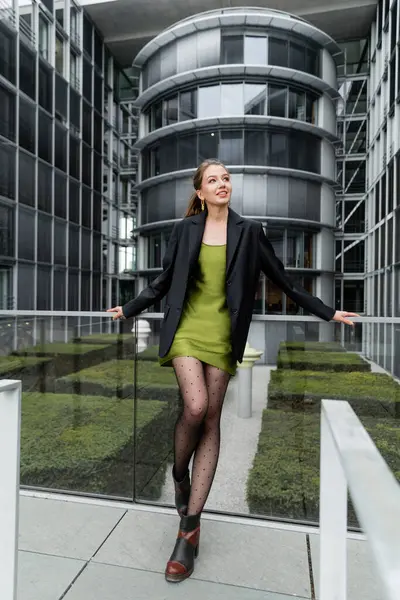 Giovane donna sorridente in giacca, abito di seta e collant a pois guardando altrove mentre in piedi a Berlino — Foto stock