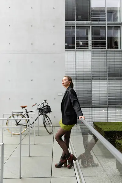 Mujer joven elegante y bonita en chaqueta negra y vestido de seda de pie cerca de la bicicleta en Berlín - foto de stock