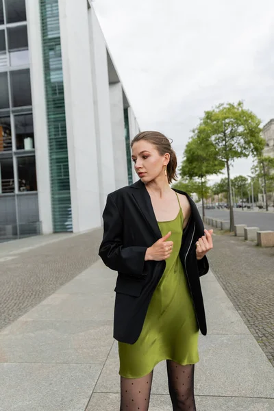 Jovem elegante em blazer preto e vestido de seda olhando para longe enquanto estava em Berlim, Alemanha — Fotografia de Stock