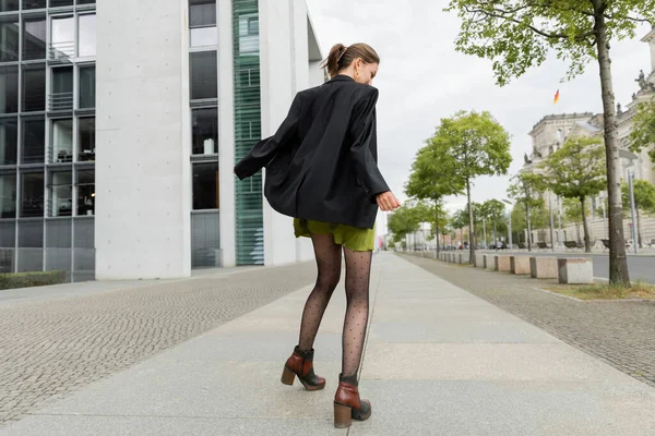 Vista lateral de la mujer joven de moda en blazer, vestido y botas caminando en la acera en Berlín - foto de stock