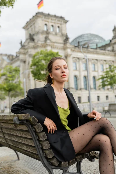 Молода жінка в куртці, шовковій сукні і колготках дивиться на камеру, сидячи на лавці в Берліні — стокове фото