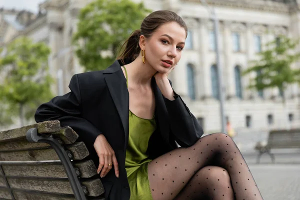 Junge Frau in Blazer, Seidenkleid und Strumpfhose, die auf einer Bank in Berlin in die Kamera blickt — Stockfoto