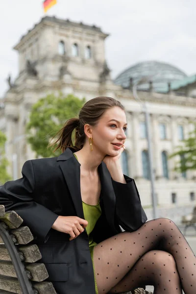 Verträumte junge Frau in Jacke, Kleid und Strumpfhose schaut weg, während sie auf einer Bank in Berlin sitzt — Stockfoto