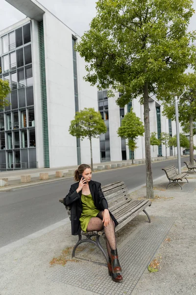 Mujer de moda en chaqueta, vestido de seda y botas hablando en smartphone en Berlín, Alemania - foto de stock