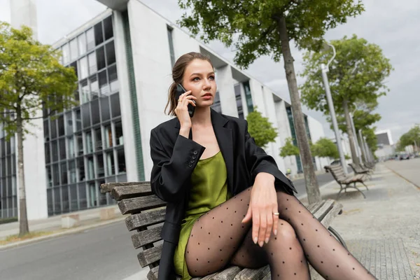 Красива волохата жінка в блістері і шовковій сукні говорить на смартфоні, сидячи на лавці в Берліні — стокове фото