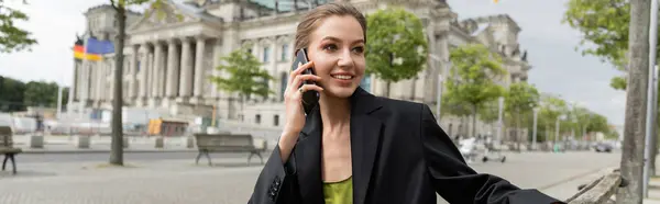 Mujer sonriente en blazer negro hablando en smartphone en la calle con Reichstag Building, pancarta - foto de stock