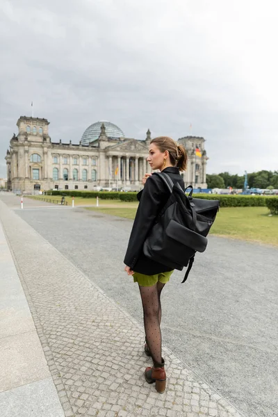 Стильна жінка в куртці і сукні тримає рюкзак під час прогулянки біля будівлі Рейхстагу в Берліні — стокове фото