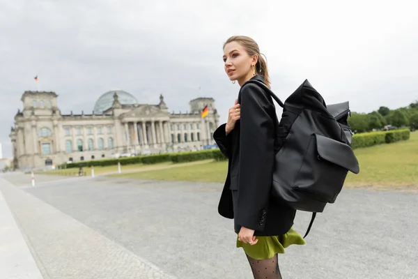 Donna alla moda in giacca e vestito che tiene lo zaino mentre cammina vicino al Reichstag Building a Berlino — Foto stock