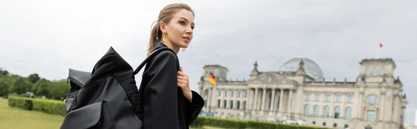 Стильна жінка в куртці і сукні тримає рюкзак під час ходьби біля будівлі Рейхстагу, банер — стокове фото