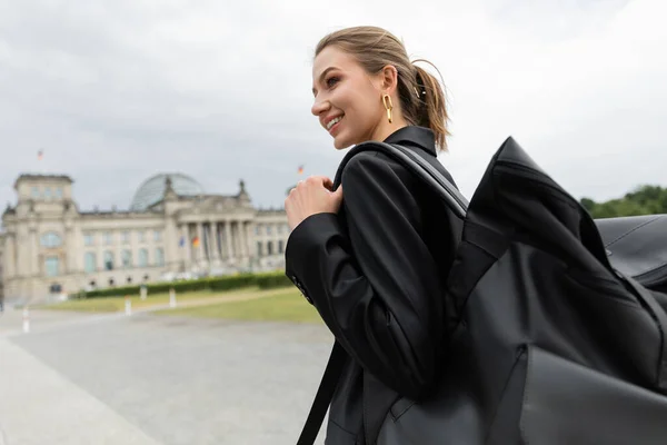Donna allegra in giacca e vestito che tiene lo zaino mentre cammina vicino al Reichstag Building a Berlino — Foto stock