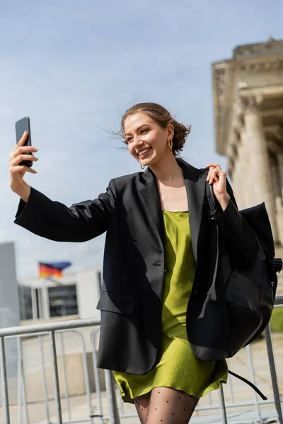Mujer alegre en chaqueta y vestido de seda sosteniendo la mochila y tomando selfie en Berlín, Alemania - foto de stock