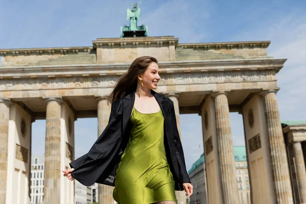 Joven alegre con chaqueta negra y vestido de seda verde cerca de la Puerta de Brandeburgo - foto de stock