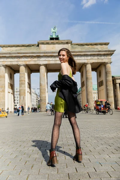 BERLÍN, ALEMANIA - 13 DE MAYO DE 2022: Mujer positiva con chaqueta negra y vestido de seda cerca de la Puerta de Brandemburgo - foto de stock