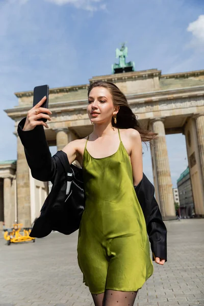 Mujer de moda en chaqueta y vestido de seda tomando selfie cerca de Brandenburg Gate en Berlín - foto de stock