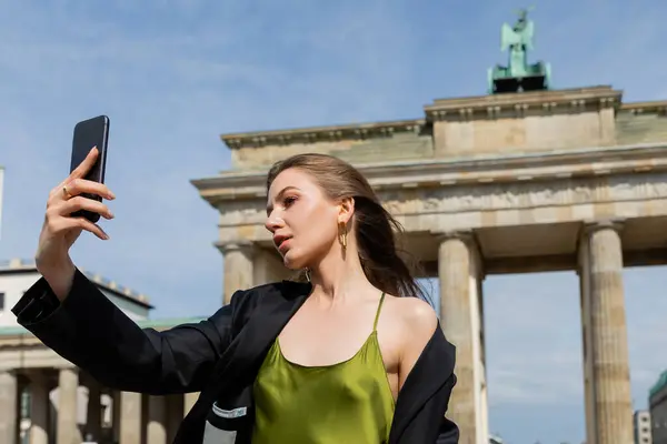 Giovane donna alla moda in giacca e abito di seta prendendo selfie vicino alla Porta di Brandeburgo a Berlino — Foto stock