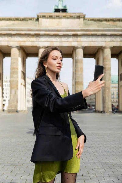 Elegante mujer de moda en chaqueta y vestido de seda tomando selfie cerca de Brandenburg Gate en Berlín - foto de stock