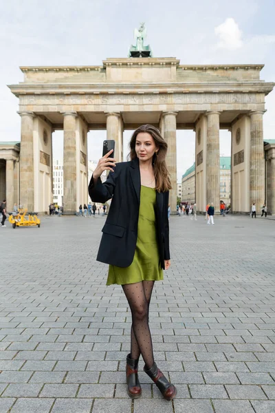 BERLÍN, ALEMANIA - 13 DE MAYO DE 2022: mujer en blazer y vestido de seda tomando selfie cerca de la Puerta de Brandenburgo - foto de stock