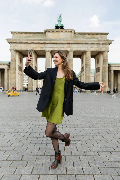 BERLÍN, ALEMANIA - 13 DE MAYO DE 2022: mujer feliz en chaqueta y vestido tomando selfie cerca de la Puerta de Brandeburgo - foto de stock