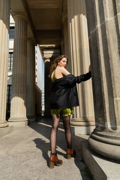 Jolie jeune femme en robe de soie tenant veste noire et debout à Berlin, Allemagne — Photo de stock