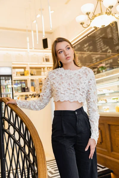 Jeune femme à la mode et aux cheveux blonds en dentelle haut et pantalon touchant rampe dans un café — Photo de stock