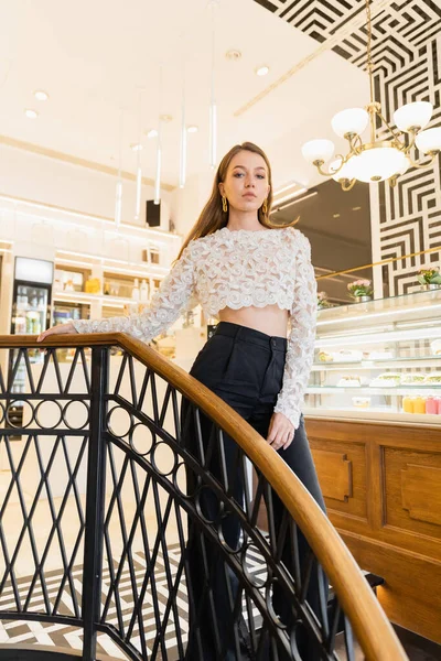 Trendy young and fair haired woman in lace top and pants standing in coffee shop — Stock Photo