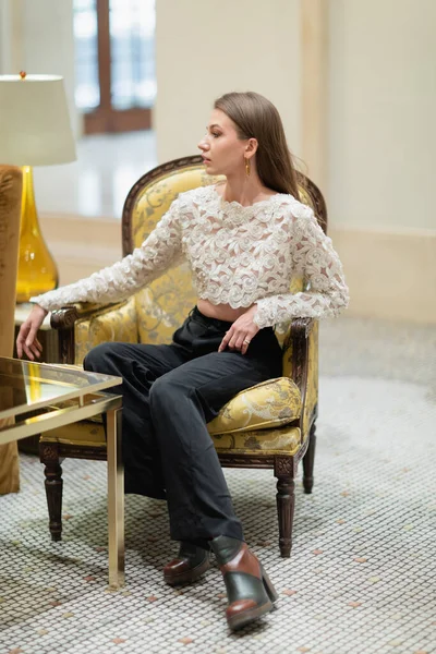 Donna dai capelli biondi in pizzo, pantaloni a vita alta e stivali che distolgono lo sguardo mentre si siede nella hall dell'hotel — Foto stock