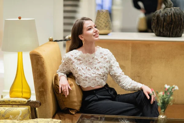 Mujer sonriente y de pelo rubio con top de encaje y pantalones sentados en el sofá en el vestíbulo del hotel de Berlín - foto de stock