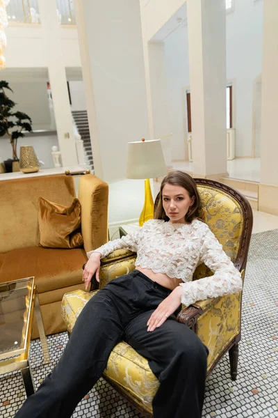 Jovem e elegante mulher no topo do laço e calças olhando para a câmera enquanto sentado na poltrona — Fotografia de Stock
