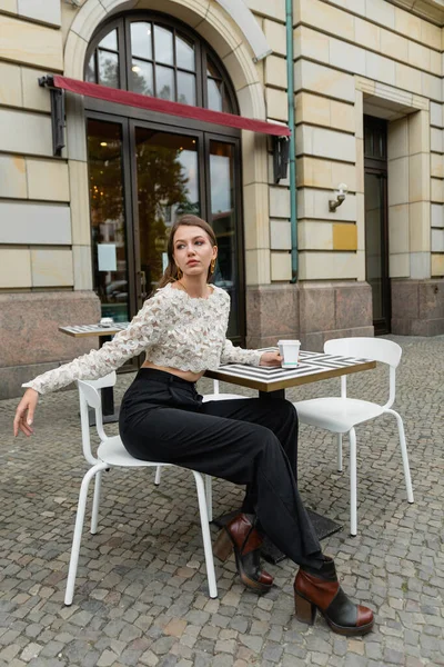 Jovem mulher no topo do laço e calças olhando embora enquanto segurando café e sentado perto de café ao ar livre — Fotografia de Stock